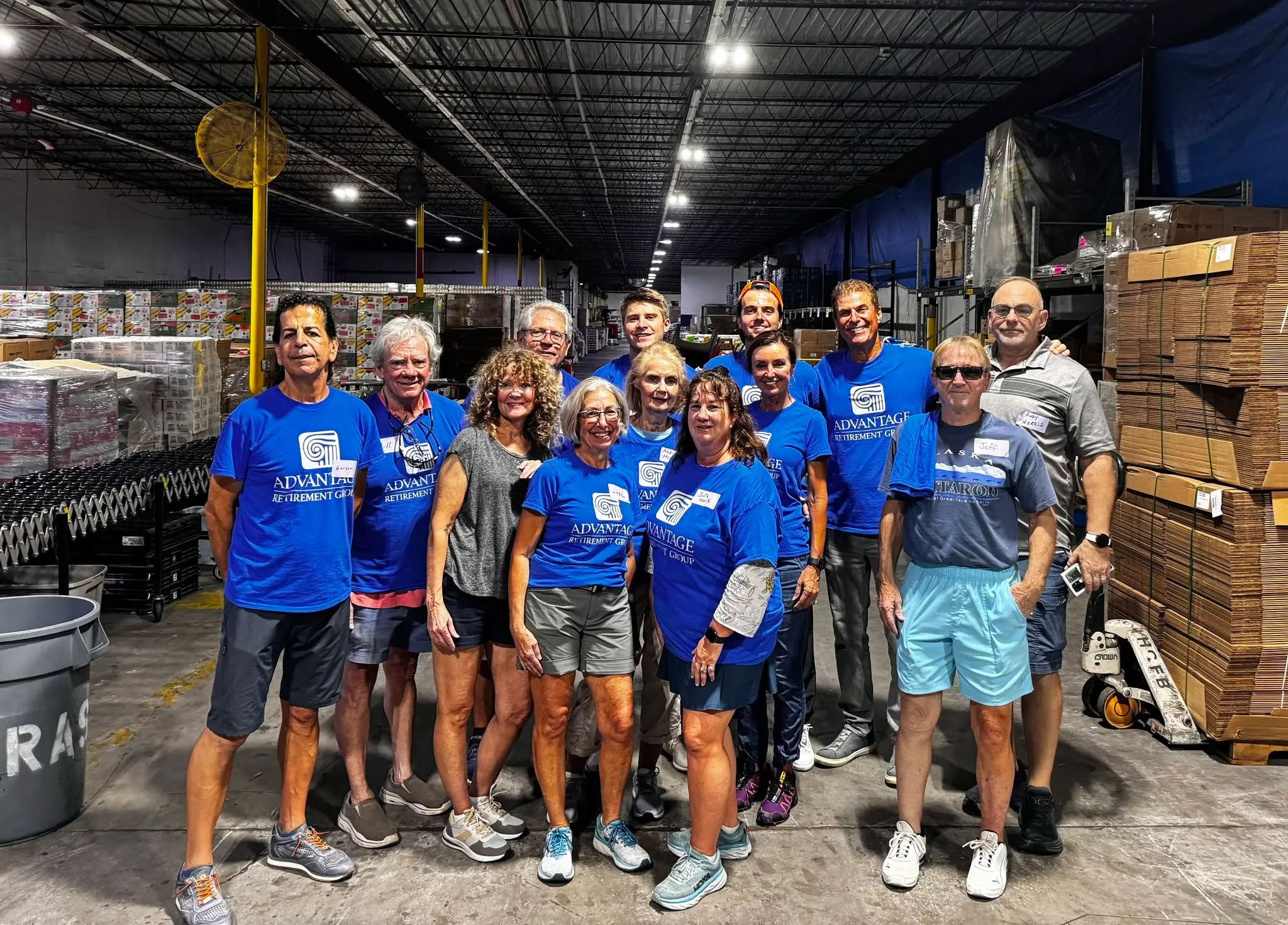 Volunteers from Advantage Wealth Partners and their clients standing together in a food bank warehouse, pallets of boxed food stacked behind them