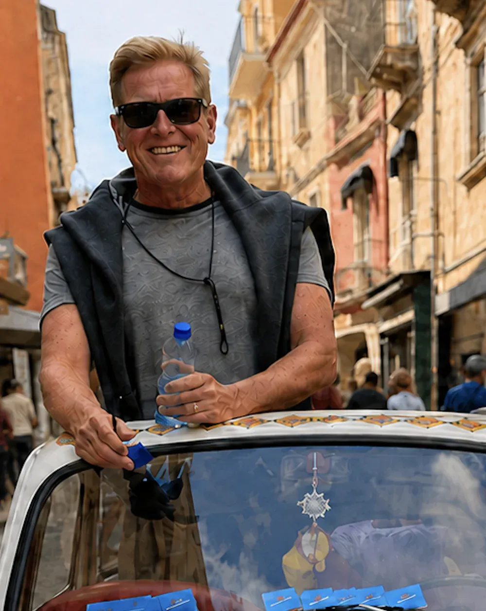 Jeff Lavender in sunglasses, smiling as he leans over the roof of a white Fiat 500 hand-painted with bright Sicilian folk-art panels, on a sunlit street of ochre stone buildings.